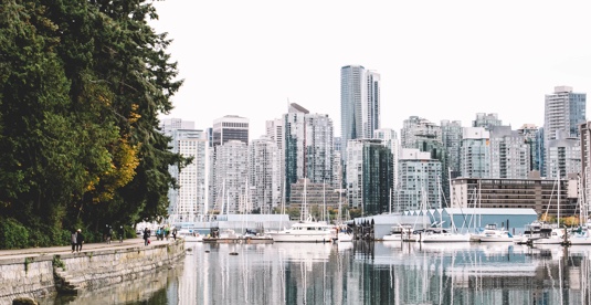 A photograph of Vancouver taken from the see wall. It shows a treed part of the seawall, with glass buildings in the background